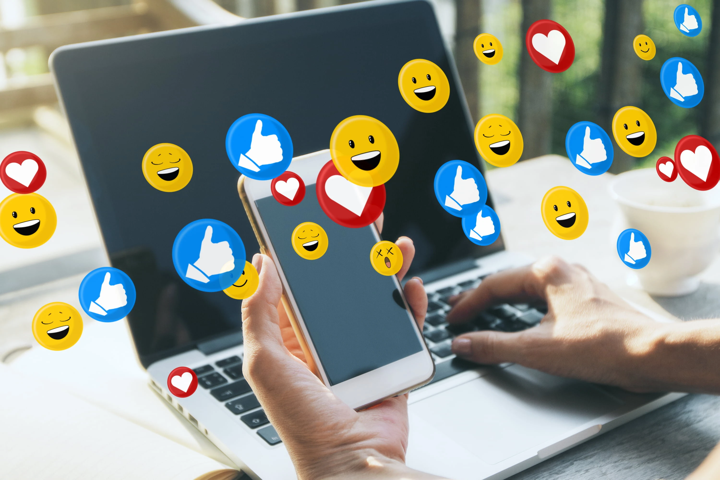 A hand holding a smartphone surrounded by floating like, heart, and emoji icons